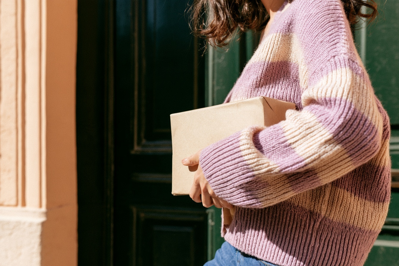A shopper in a lilac striped knit carrying a parcel beside a green front door.